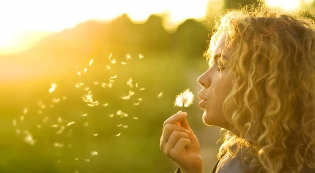 Una joven soplando una flor blanca de diente de león, con los ojos cerrados mientras las semillas de la flor vuelan en el aire